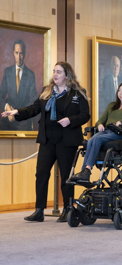 Accessible tours through Australian Parliament House Young female tour guide shows a young woman in a mobility chair through portraits hung on the walls inside Australian Parliament House.