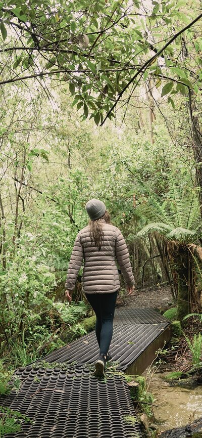 Tidbinbilla Cascade Trail | © We Are Explorers Image of a woman in a light puffer jacket and hat walking the Cascade Trail through trees in Tidbinbilla Nature Reserve. | © We Are Explorers