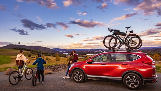 Explore outdoors at the National Arboretum Canberra Two young kids on bikes and a couple lean against a red car bonnet overlooking the view of Black Mountain Tower at the National Arboretum Canberra