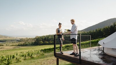 A couple pouring wine on a deck in front of a vineyard.