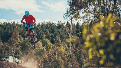 A man in a red shirt and blue shorts on a black bike jumping on a outdoor trail with a forest in the background.