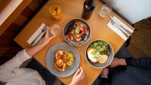 Three different colourful brunch dishes on a wooden table with two people sitting nearby.