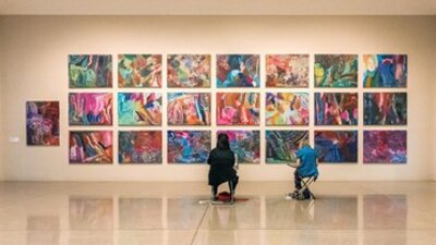 Two visitors to the gallery sit in front of a large colourful artwork.