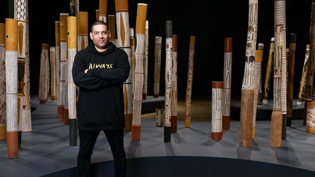 Tony Albert (Girramay/Yidinji/Kuku-Yalanji peoples) in The Aboriginal Memorial, 1987–88 A man standing in a gallery space with indigenous poles