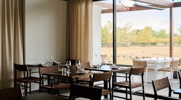 Modern restaurant interior with wooden tables set for dining, large windows, and a view of trees outside