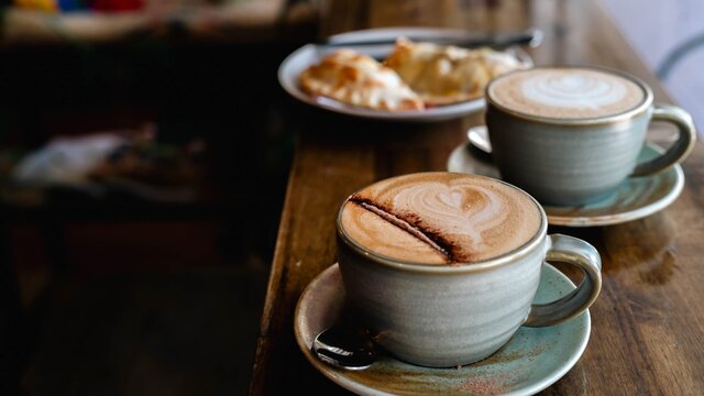 Two hot drinks in mugs with food plated in the background