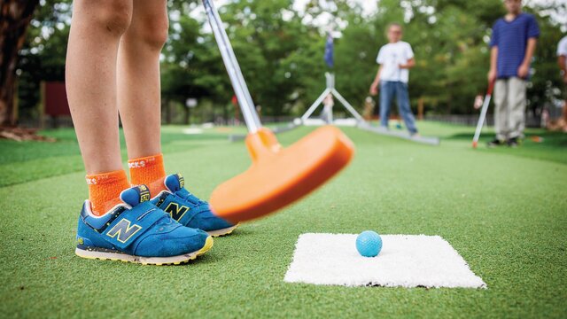 A low ground perspective show a kid getting ready to put a golf ball into a whole under a mini replica Australian Parliament House.